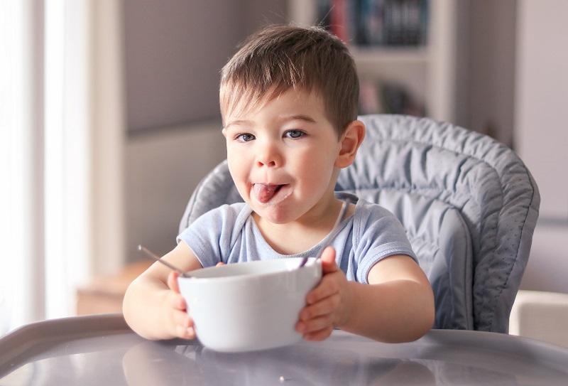 Adorable thankful little baby boy with smeared face and tongue out just finished his tasty meal and hold white bowl looking at camera wanting more sitting at high feeding chair. Child nutrition concept. 